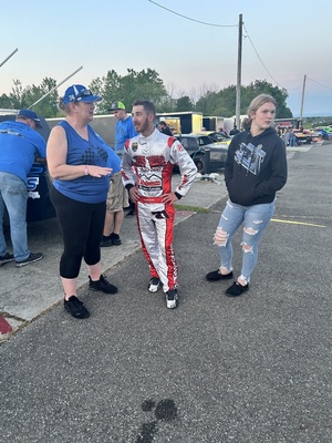 Three people, including a race car driver in suit, stand outdoors.